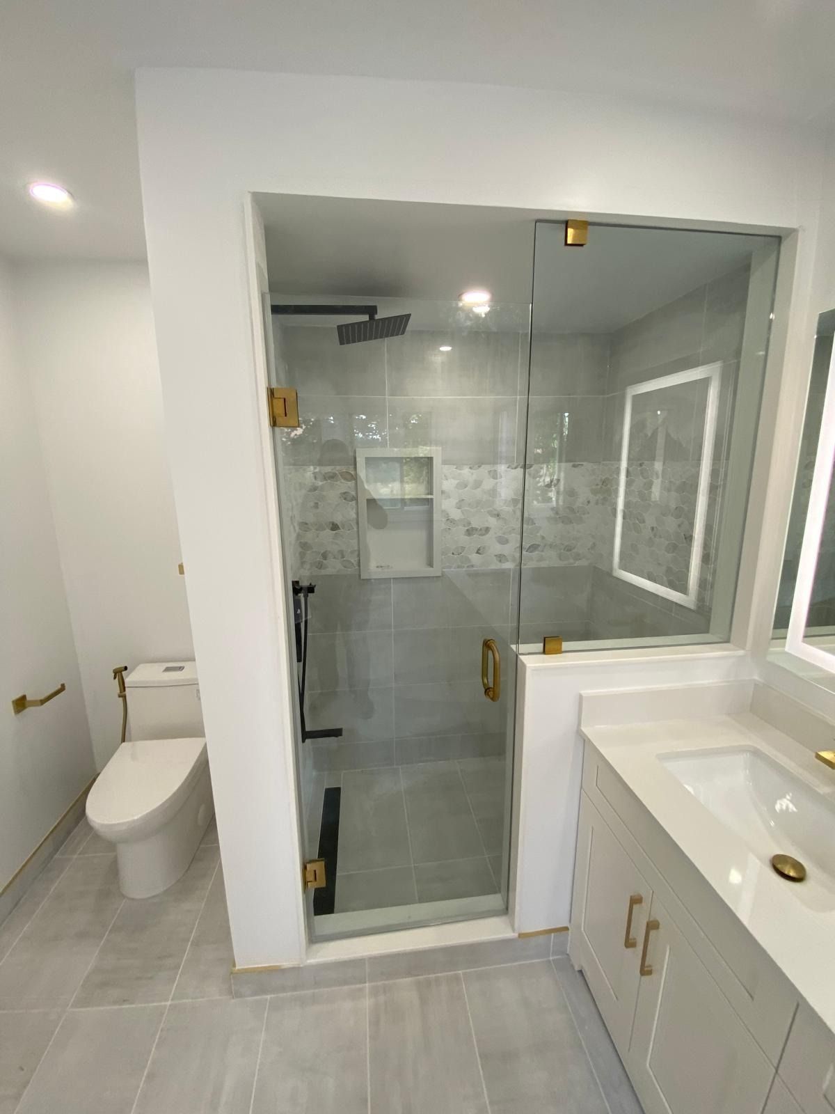 Shower with frameless glass, brass hinges and handle, a black rain head, a marble mosaic band and a lit mirror